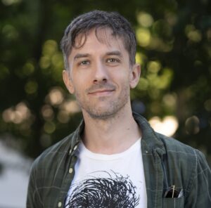 A man with short brown hair and light stubble smiles slightly at the camera. He wears a green plaid shirt over a white T-shirt with a black abstract design. The background is blurred greenery.