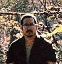 A person with glasses and a mustache stands outdoors, wearing a dark shirt with trees and autumn foliage in the background.