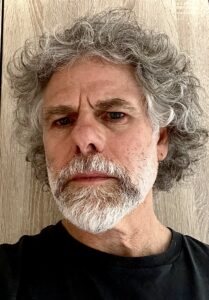 A man with curly gray hair and a beard looks into the camera with a serious expression, standing in front of a light wooden background and wearing a black shirt.