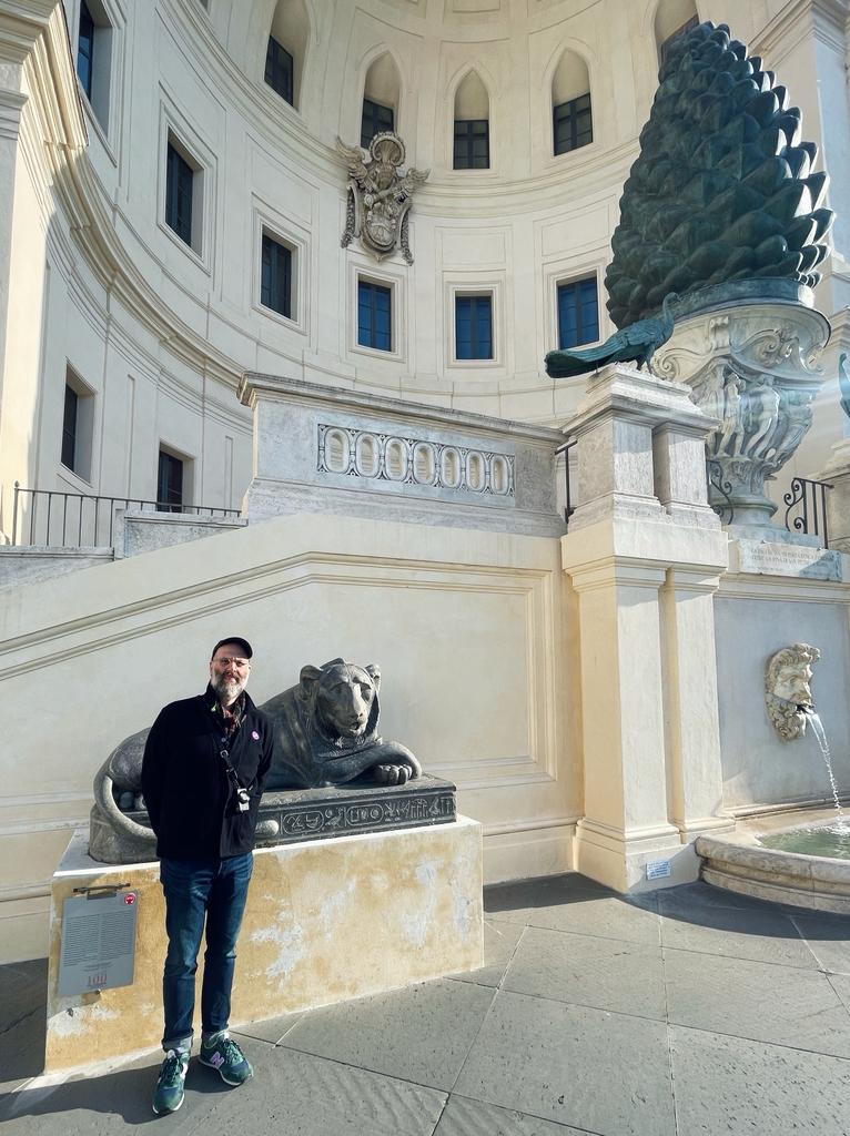 A person stands next to a stone lion statue in a sunlit courtyard with cream-colored walls, arched windows, and a large pine cone sculpture atop a fountain structure.